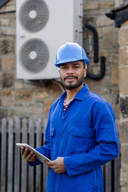 Male construction worker standing in front of a home where he is installing an air source heat pump on the side of a home in the North East of England, which is out of focus. The house is aiming to be sustainable. He is smiling, looking at the camera while holding a digital tablet.