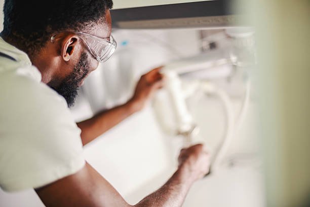 Professional black man plumber wearing safety glasses and installing a sophisticated water filter system under a kitchen sink, ensuring clean water and home maintenance